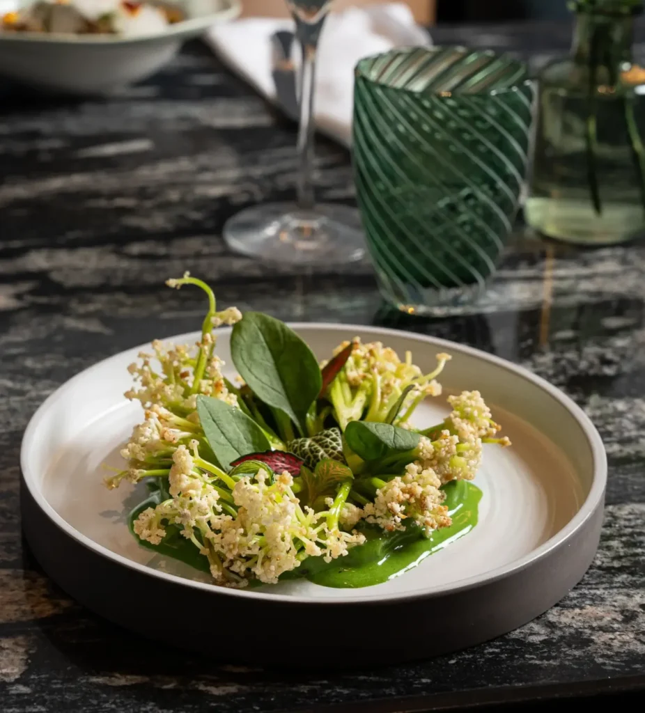 A modern dish at Virens Restaurant is garnished with edible green leaves and white blossoms, presented on a white plate with green sauce, set on a marble table next to a green glass.