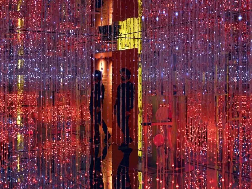 People stand among hanging strings of red and white LED lights in an immersive, mirrored art installation at the White Rabbit Museum.