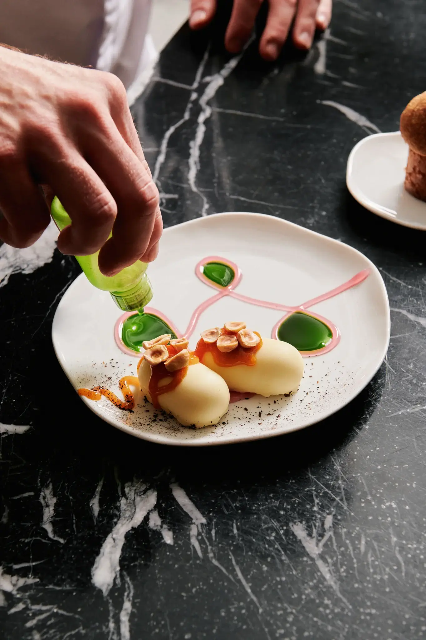 Die Hand eines Kochs verziert ein plattiertes, pflanzenbasiertes Dessert mit grüner Soße auf einer weißen Schale mit kunstvollen rosa und grünen Mustern, die auf einer schwarzen Marmorarbeitsplatte steht.