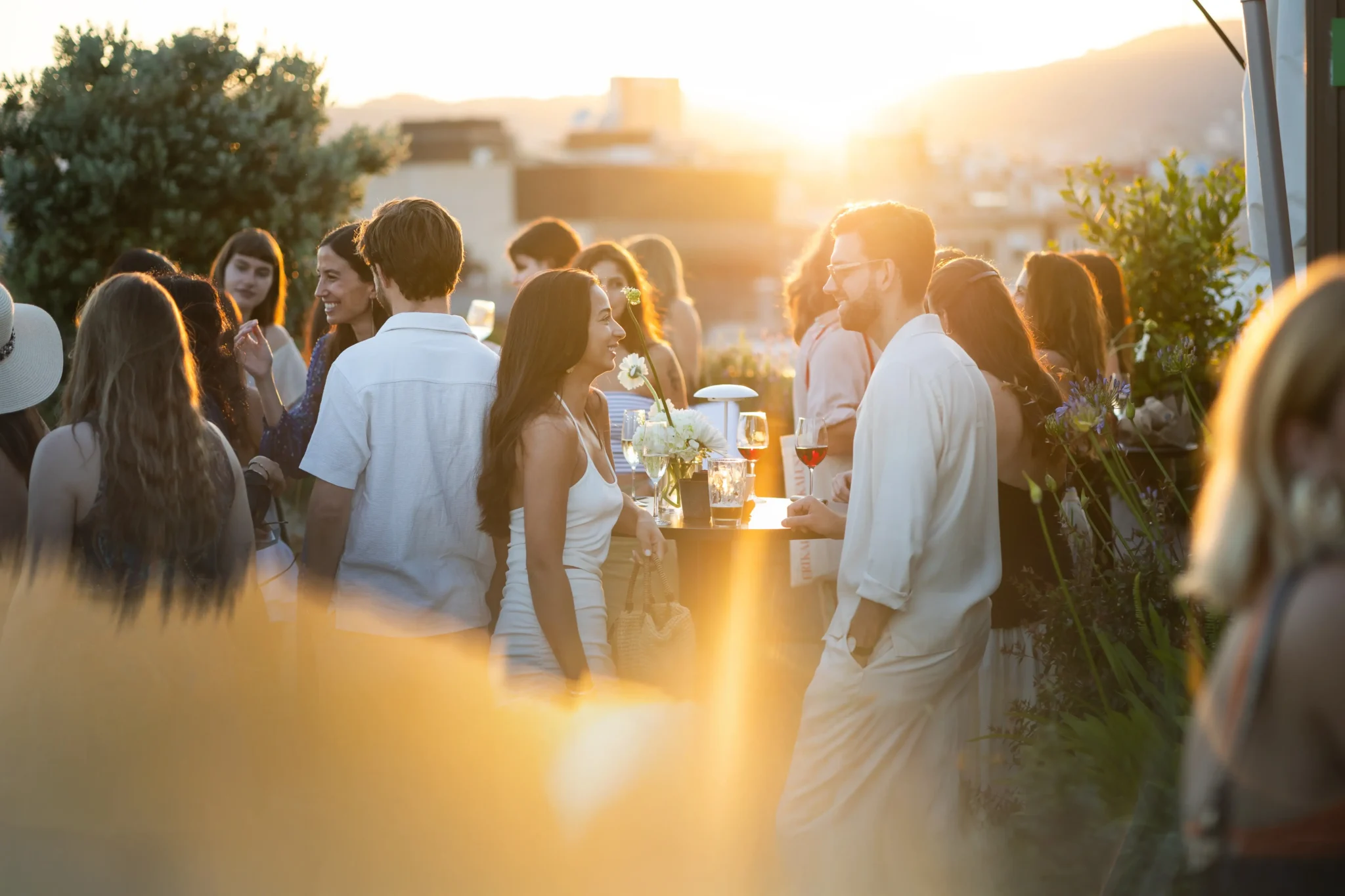 Un grup de persones socialitzen a l'aire lliure a la posta de sol durant actes socials, drets al voltant de taules amb begudes. L'entorn sembla relaxat, amb una càlida llum del sol i vegetació al fons.
