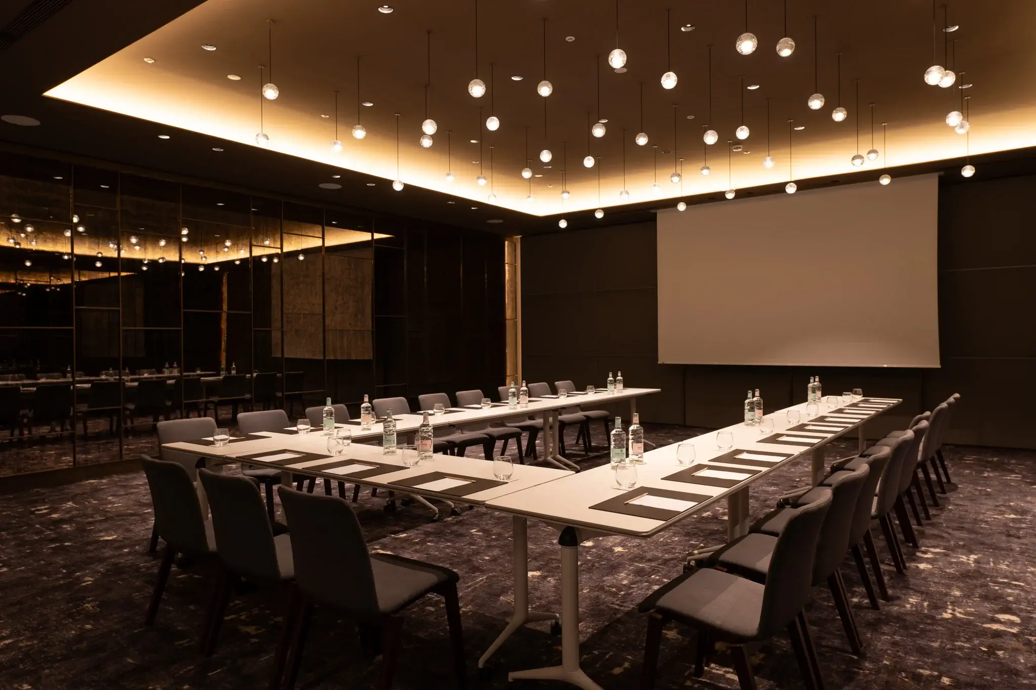 A modern conference room for meetings or events, featuring U-shaped tables, chairs, bottled water, notepads, a large projection screen, and ceiling lights.