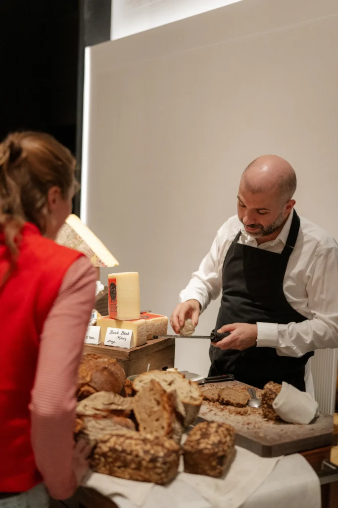 A man in an apron slices bread at a table with various types of bread and cheese, while a woman in a red jacket stands nearby, savoring flavors that evoke the charm of culinary destinations.