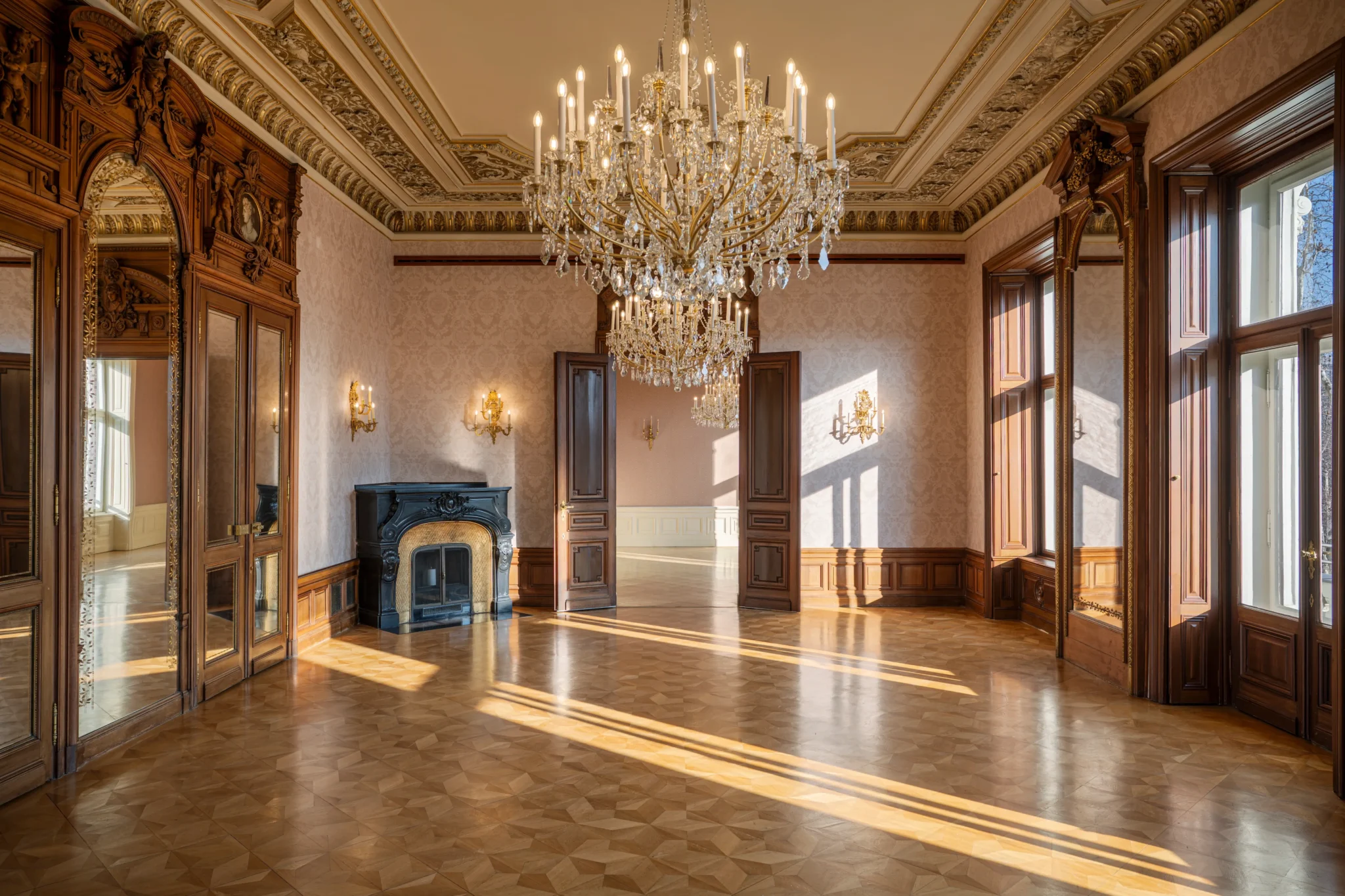 Verschnörkelter, leerer Raum mit großen Fenstern, Parkettboden, Kristalllüstern, Holzvertäfelung, Wandspiegeln und einem dekorativen schwarzen Kamin, der von natürlichem Sonnenlicht beleuchtet wird.