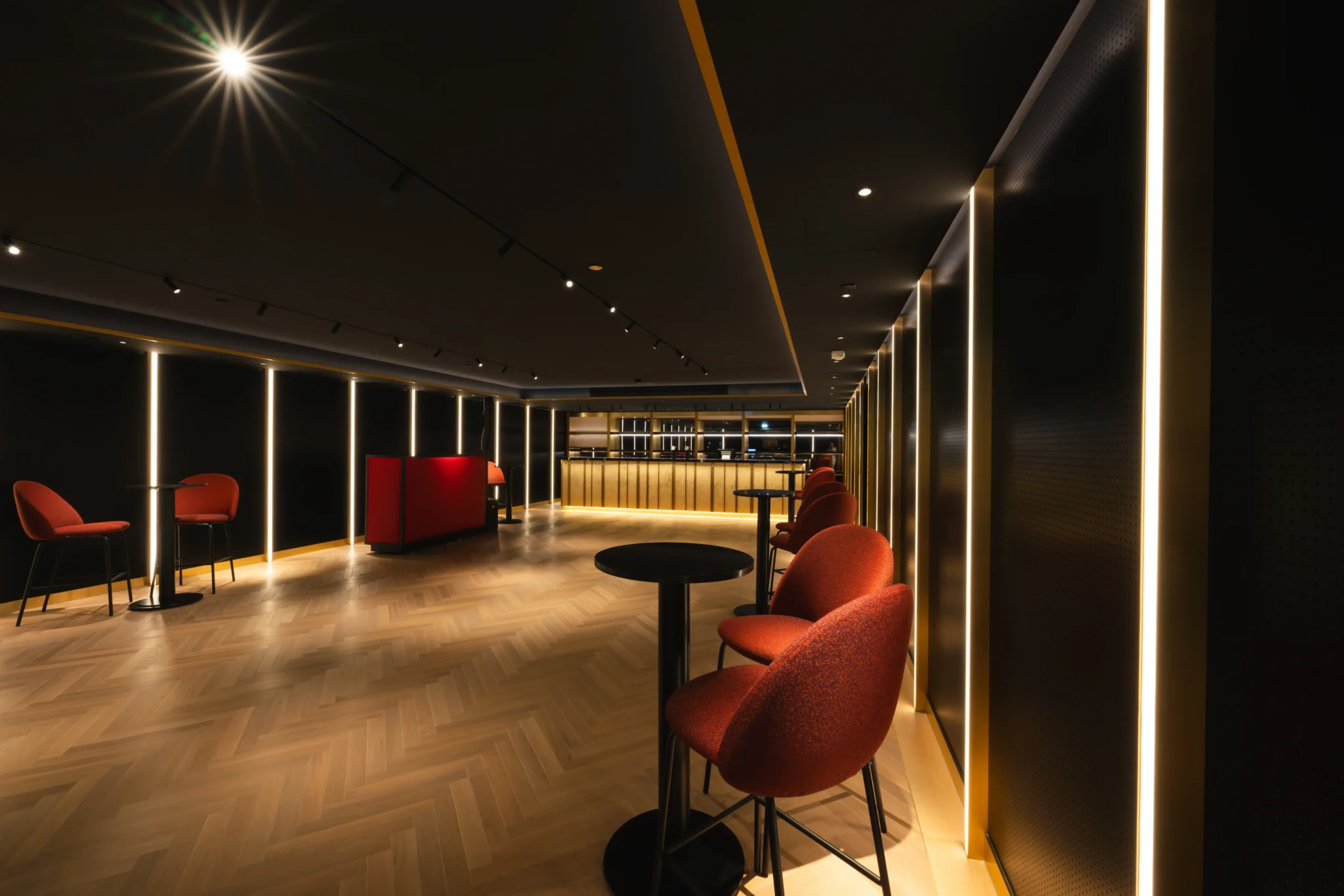 Eine moderne Lounge mit warmer Beleuchtung, roten Stühlen, kleinen Tischen und einem Holzboden, mit einem Barbereich im Hintergrund.