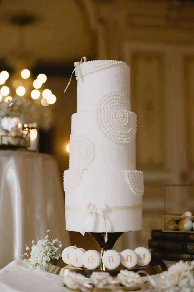 A three-tiered white wedding cake decorated with pearl-like beads sits on a stand; white iced cookies spelling “Vienna” are arranged at its base, making it the perfect centerpiece for special events or gatherings, such as a memorable wedding meeting.