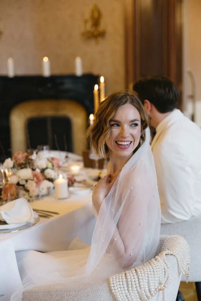 Eine Braut in einem weißen Kleid und Schleier lächelt, während sie an einem geschmückten Tisch neben einem Mann in einem weißen Anzug bei einer formellen Veranstaltung im Haus sitzt.