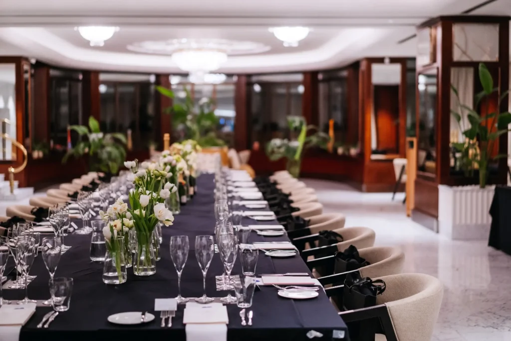 A long dining table set with a black tablecloth, glassware, plates, and flower arrangements awaits social gatherings or weddings in an elegant, well-lit banquet hall.