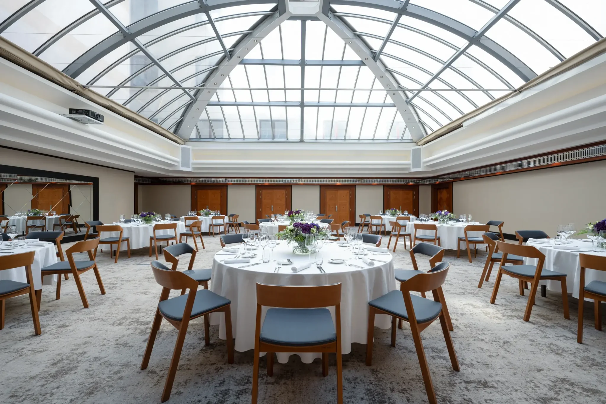 V prostorné místnosti s proskleným stropem jsou kulaté stoly s bílými ubrusy, židlemi a květinovými dekoracemi připravené pro slavnostní události nebo bankety.