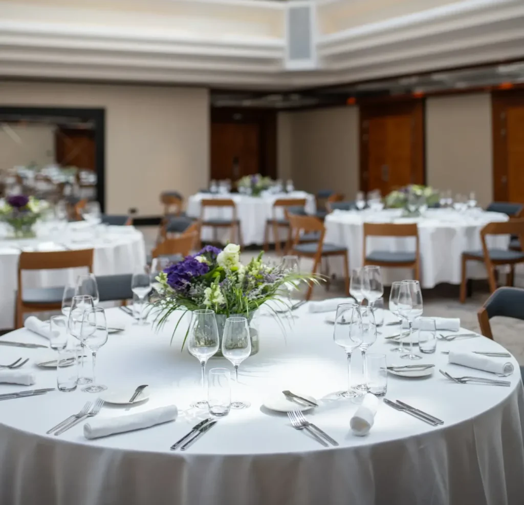 Runde Banketttische mit weißen Tischtüchern, Gläsern, Besteck und Blumenschmuck schaffen eine einladende Atmosphäre für Tagungen und Veranstaltungen in einem geräumigen, hellen Veranstaltungssaal.