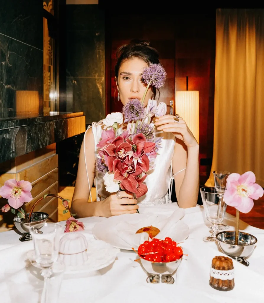 A woman in a white dress sits at a set dining table during a wedding, holding a bouquet of flowers in front of her face, with various desserts and glassware arranged around her.