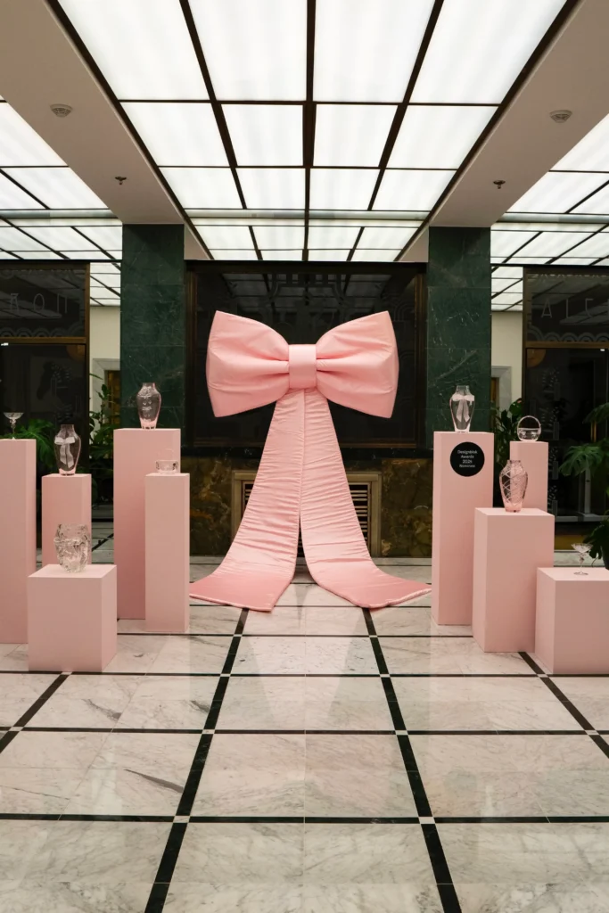A large pink bow is displayed on a wall, surrounded by glass vases on pink pedestals in a brightly lit, marble-floored room—an artful showcase of Inspired Spaces.