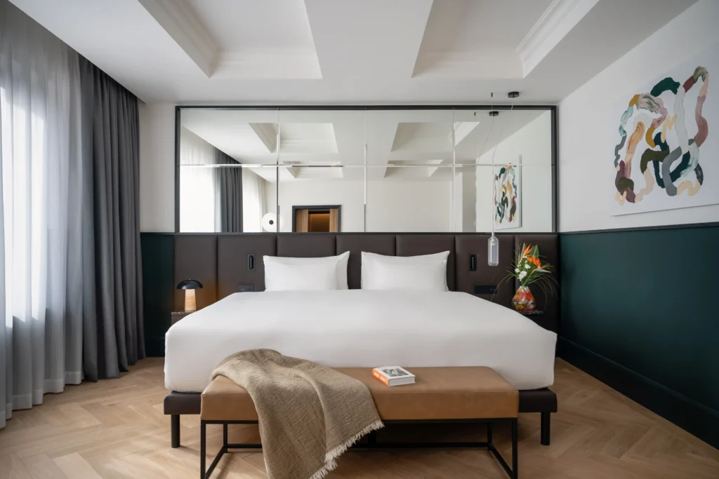 Modern hotel room at Almanac Alcron Prague with a large bed, brown bench, book and blanket, floor-to-ceiling curtains, abstract wall art, and a striking wall mirror behind the bed.