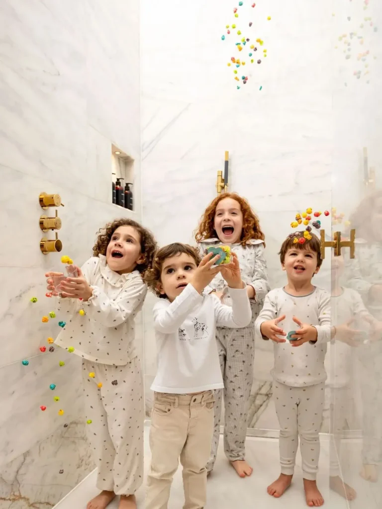 Four young children in pajamas stand in a marble shower, reaching up and smiling as colorful toy balls fall around them, celebrating the joy of a Longer August.