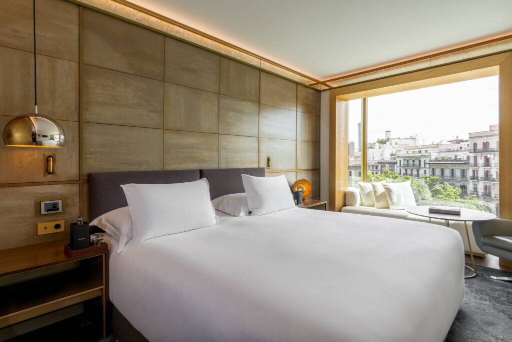 Una habitació d'hotel moderna de l'Almanac Barcelona disposa d'un llit gran, dues tauletes de nit, un seient a la finestra i unes vistes impressionants dels edificis de la ciutat a través d'una finestra espaiosa.