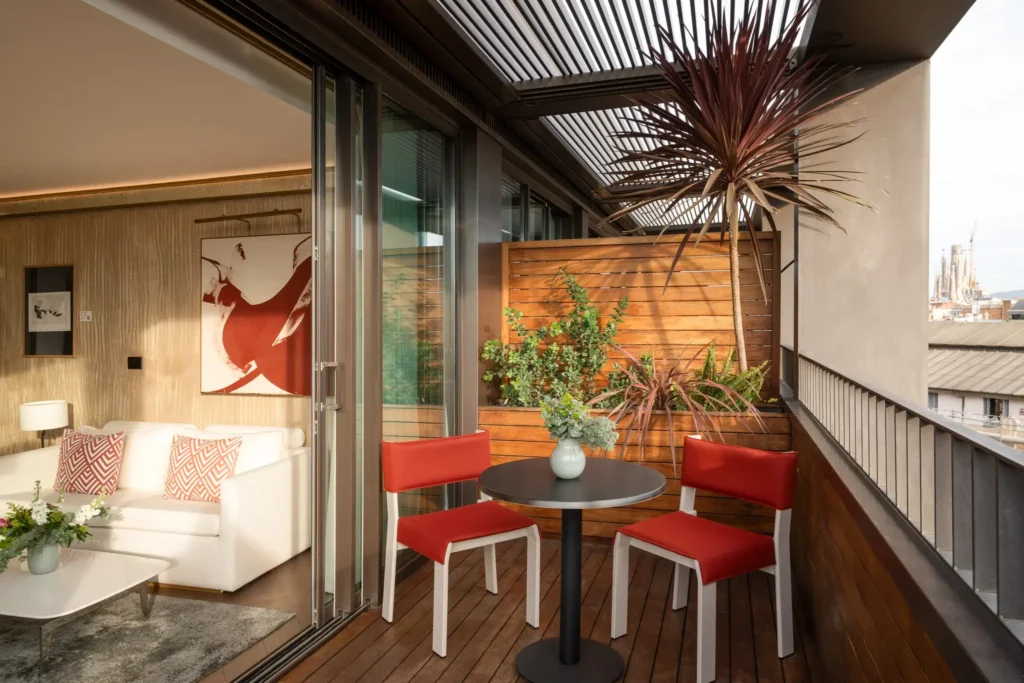 A modern balcony at Almanac Barcelona features two red chairs, a round table with a vase, potted plants, and offers a view into a living room with a white sofa and abstract art.