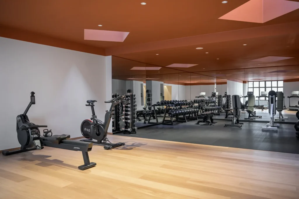 Moderno gimnasio con bicicletas estáticas, pesas libres, bancos y equipos de entrenamiento de fuerza, con una pared de espejos y detalles naranjas en el techo.