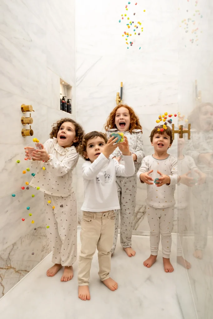 Quatre nens petits en pijama estan drets en una dutxa de marbre, agafant joguines de ventosa de colors enganxades a les parets i flotant a l'aire a l'Almanac Barcelona, somrient i jugant junts.