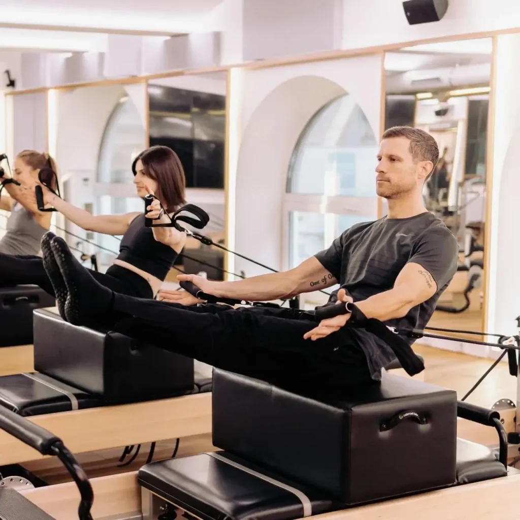 Three people exercise on Pilates reformer machines, holding resistance bands and balancing with legs raised in an X-Treme Pilates fitness studio lined with mirrors.