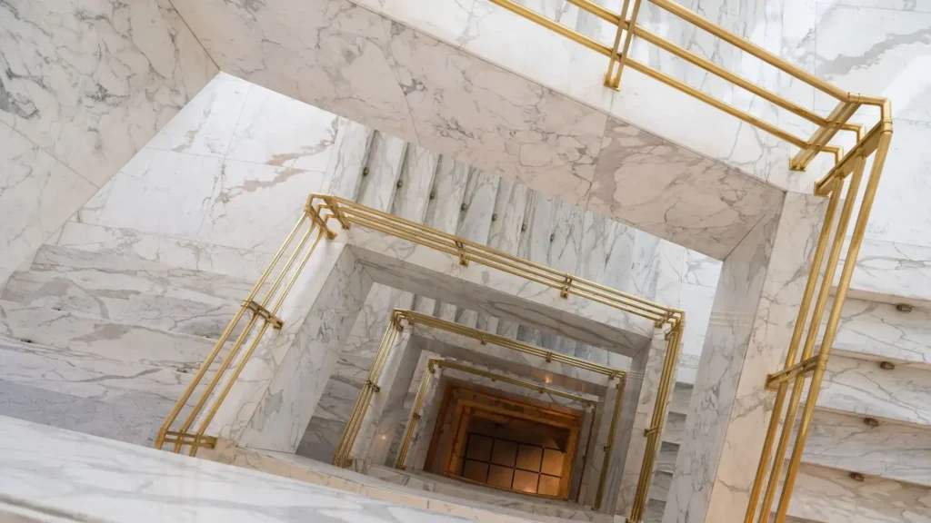 Looking down a spiral staircase with white marble walls and gold railings, forming geometric patterns—perfect inspiration for any Sales Manager seeking elegance in design.