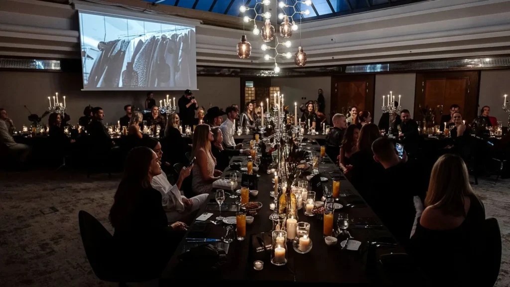 A group of people sit around a long, candle-lit banqueting table in a dimly lit room with a large screen displaying an image of clothing racks.