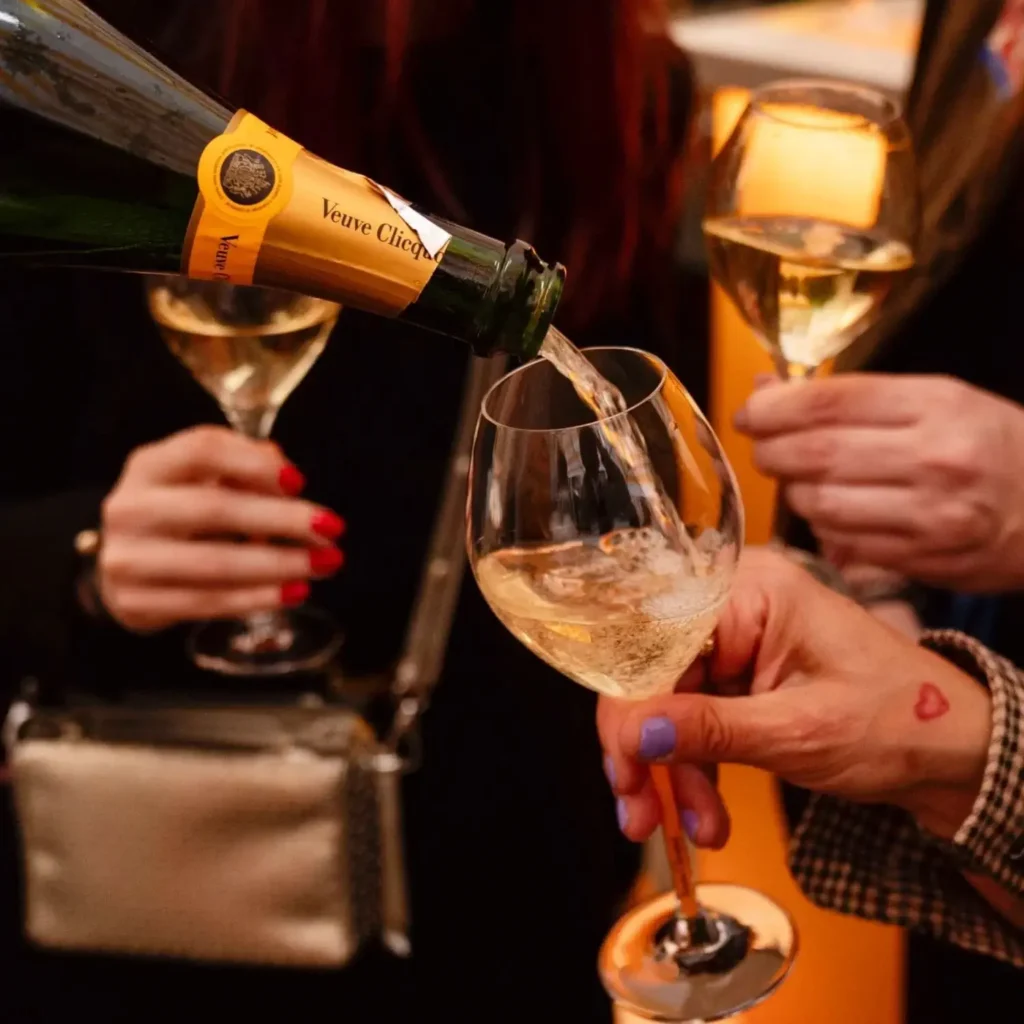 Eine Flasche Veuve Clicquot Champagner wird in ein Glas gegossen, während zwei Personen im Hintergrund gefüllte Champagnergläser halten.