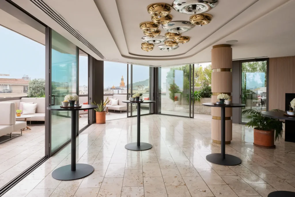 Moderner Veranstaltungsraum mit großen Fenstern, runden Cocktailtischen, Marmorfußboden und Zugang zu einer Außenterrasse - perfekt für Meetings und Veranstaltungen mit Blick auf die Stadt.