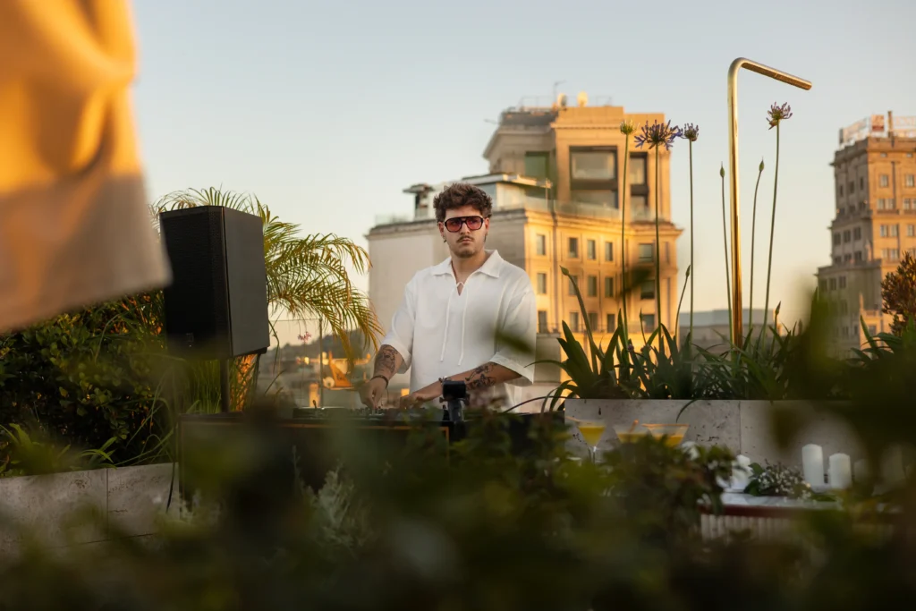 Un hombre con gafas de sol y camisa blanca está de pie junto a una mesa de DJ en una azotea con edificios urbanos y plantas de fondo.