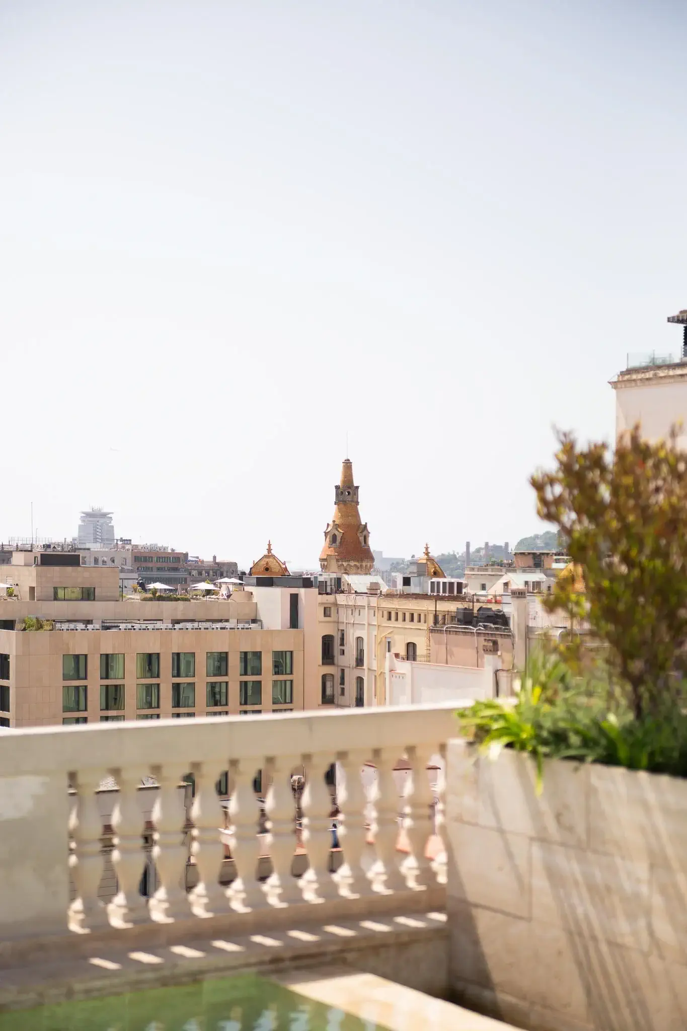 Vista des de la teulada d'un paisatge urbà amb edificis històrics, inclosa una torre prominent, que es veu darrere d'una balustrada de pedra i plantes en primer pla: un lloc ideal per relaxar-se i gaudir de l'encant de Terrassa després d'un bany a la piscina.
