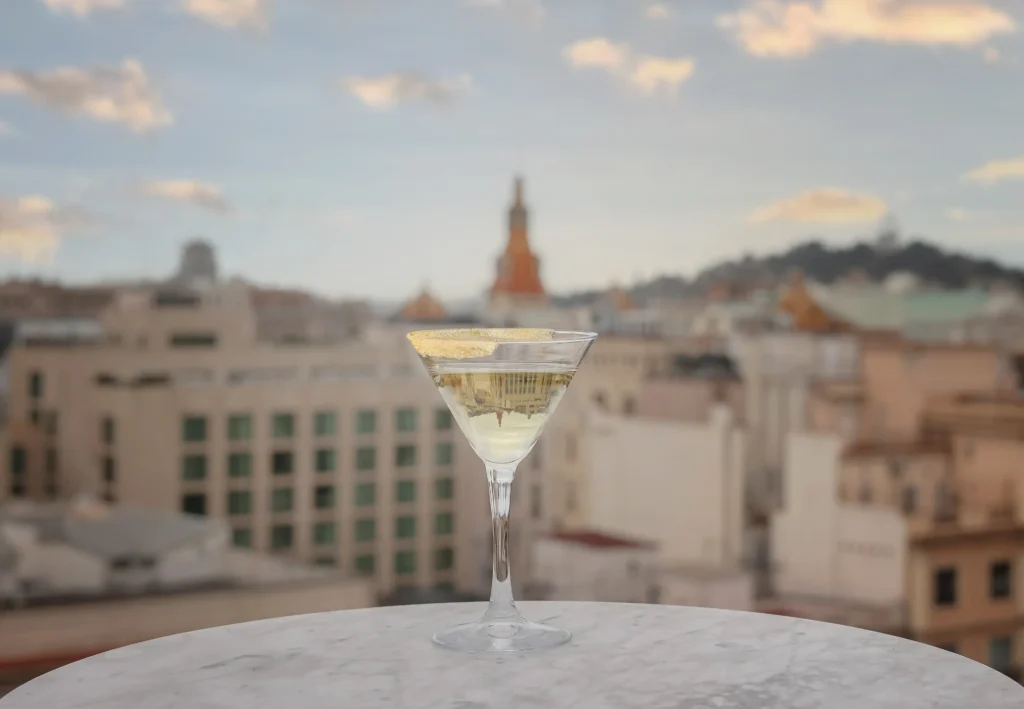 Una copa de martini plena d'una beguda clara es troba sobre una taula de marbre, amb un paisatge urbà borrós i un cel ennuvolat al fons.