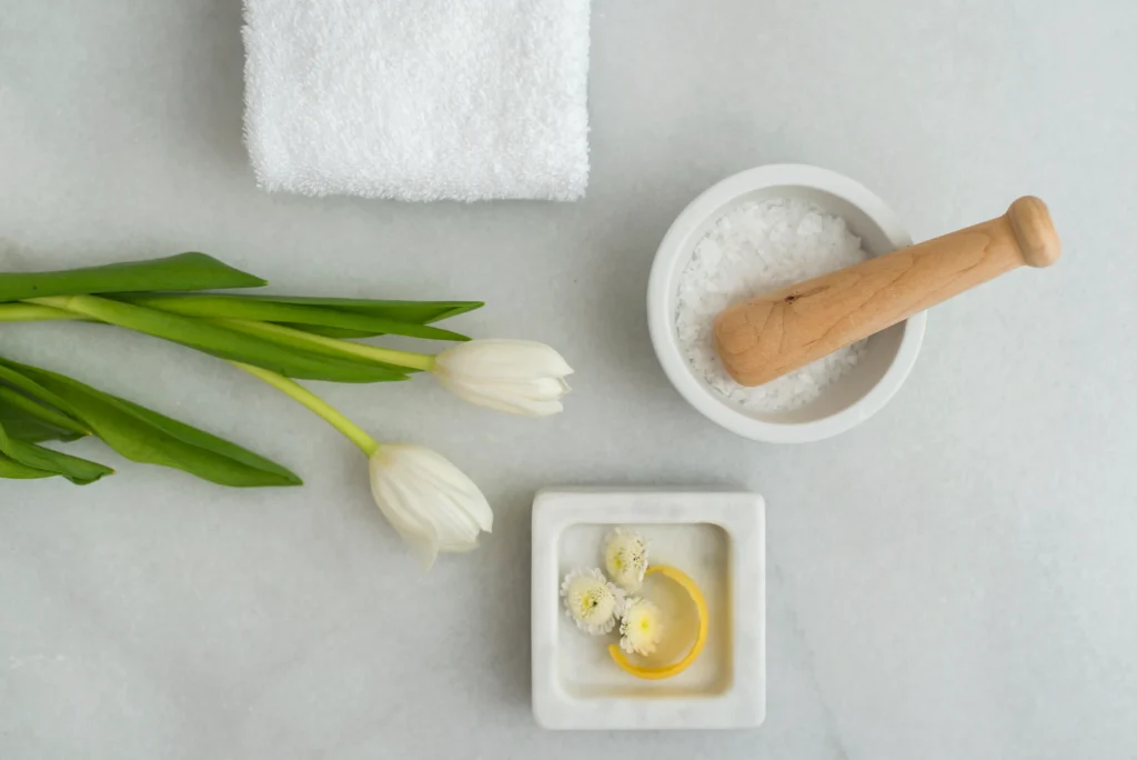 A folded white towel, three white tulips, a bowl of bath salts with a wooden pestle, and a small dish with yellow oil and white flowers arranged on a light surface.
