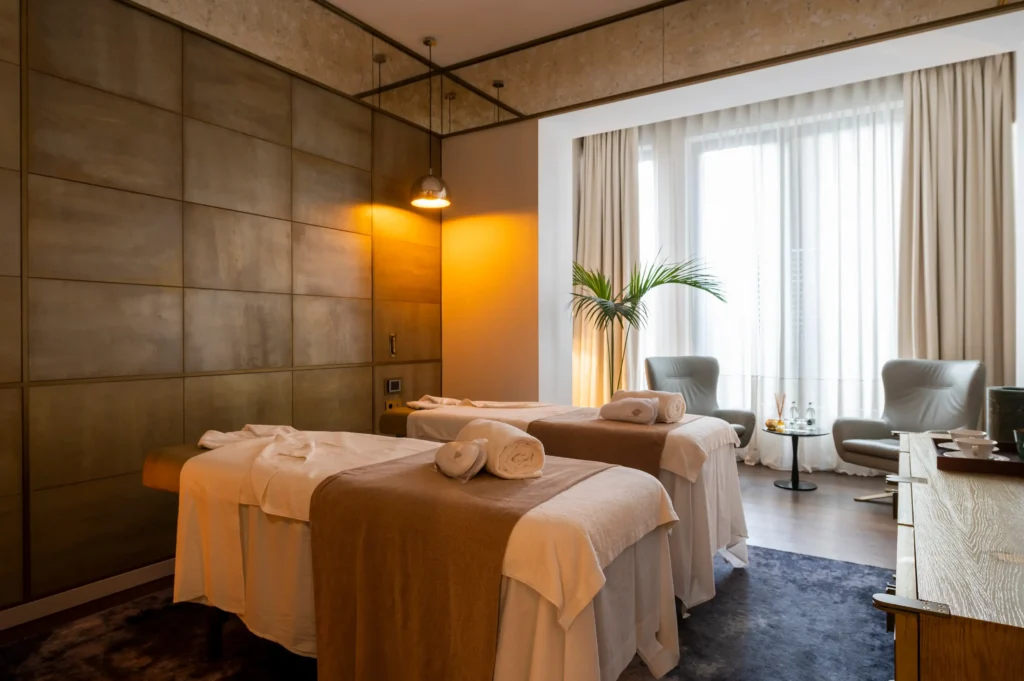 A spa room with two massage tables covered in white towels, a large window with sheer curtains, two chairs, a small table, and a potted plant.
