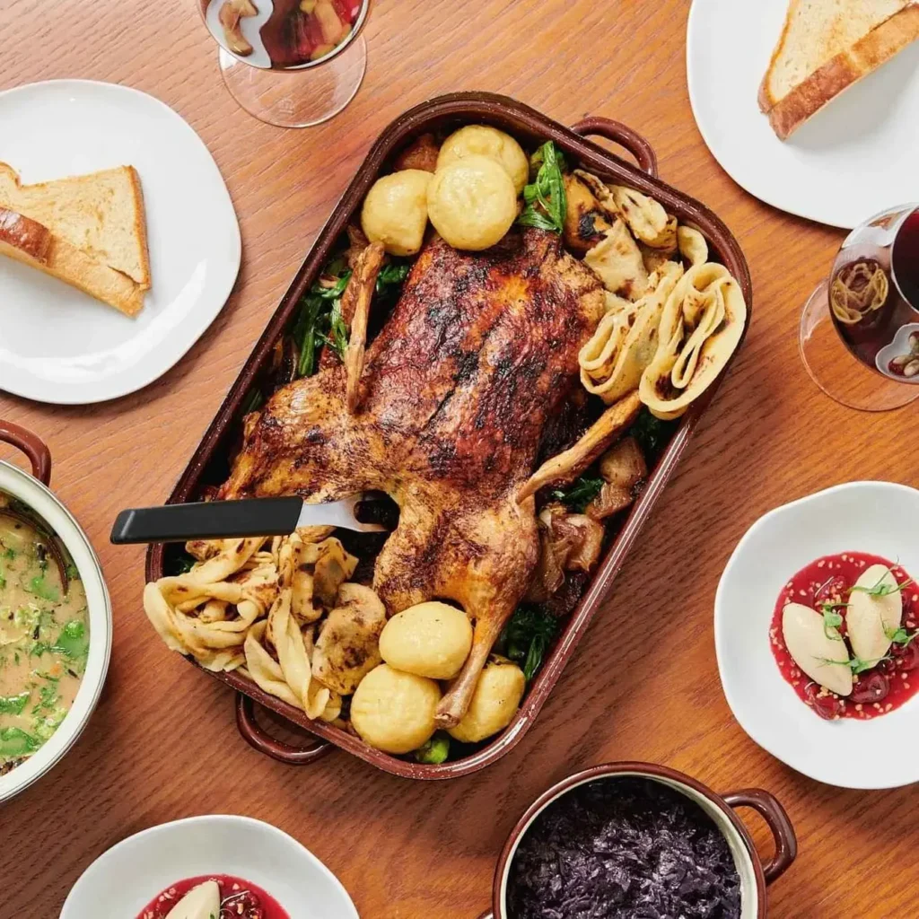 Eine gebratene Ente mit Kartoffeln und Gemüse wird in einer Auflaufform serviert, umgeben von Tellern mit Brot, Soße, Rotkohl, Suppe und Gläsern mit Rotwein auf einem Holztisch.