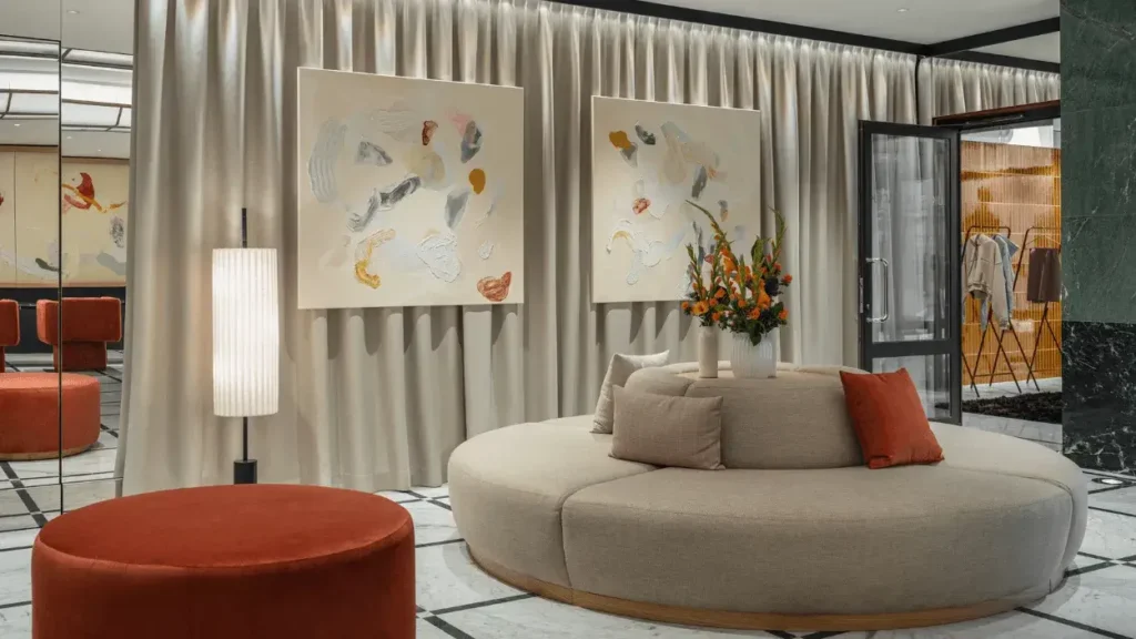 Modern lounge area ideal for a Marketing Coordinator, featuring a round beige sofa, orange ottoman, tall lamp, abstract art on curtained wall, and glass doors leading to a clothing display area.