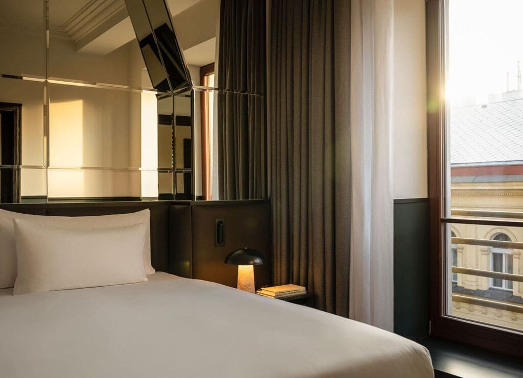 A neatly made bed with white linens, a bedside table with a lamp and book, large windows with curtains, and sunlight streaming into a modern hotel room—perfect for relaxing during our exclusive Black Friday deals.