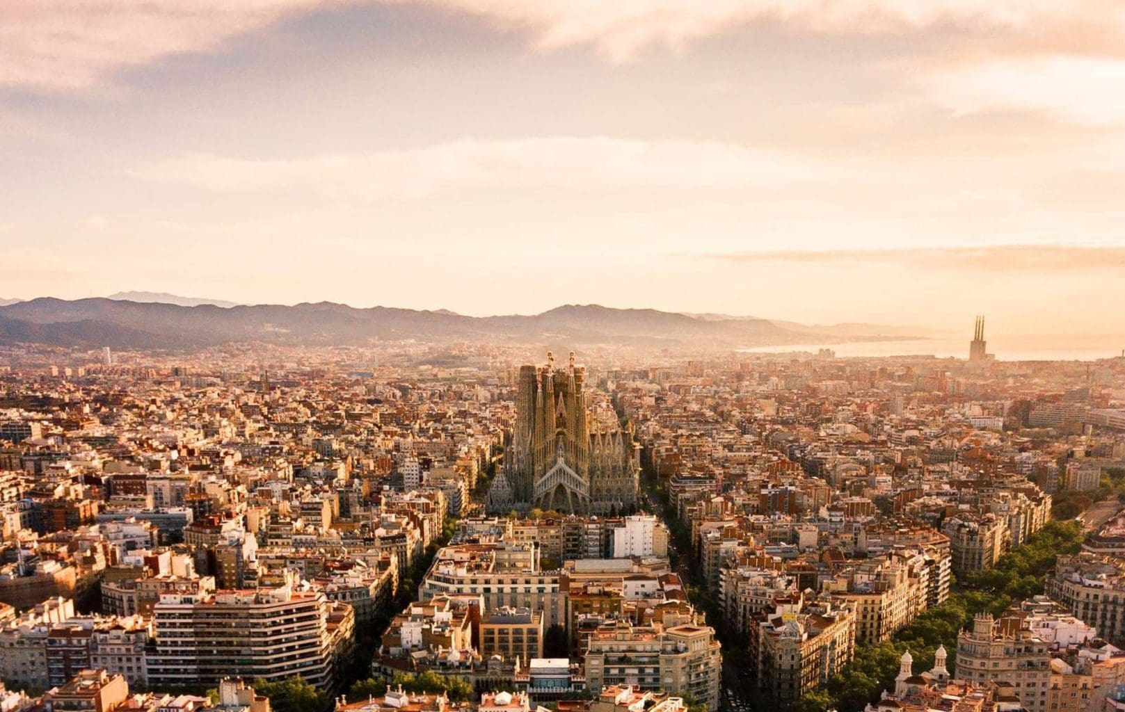 Aerial view of Barcelona at sunset, featuring the Sagrada Família basilica and the city’s grid-patterned streets, as seen from Azimuth Rooftop Bar, with distant mountains completing the stunning panorama.