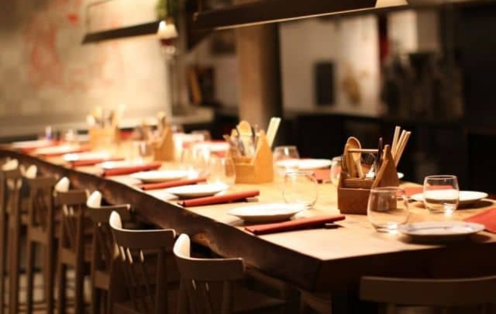 Una llarga taula de menjador de fusta amb plats, gots, tovallons i coberts, envoltada de cadires de fusta en un restaurant amb una llum tènue.