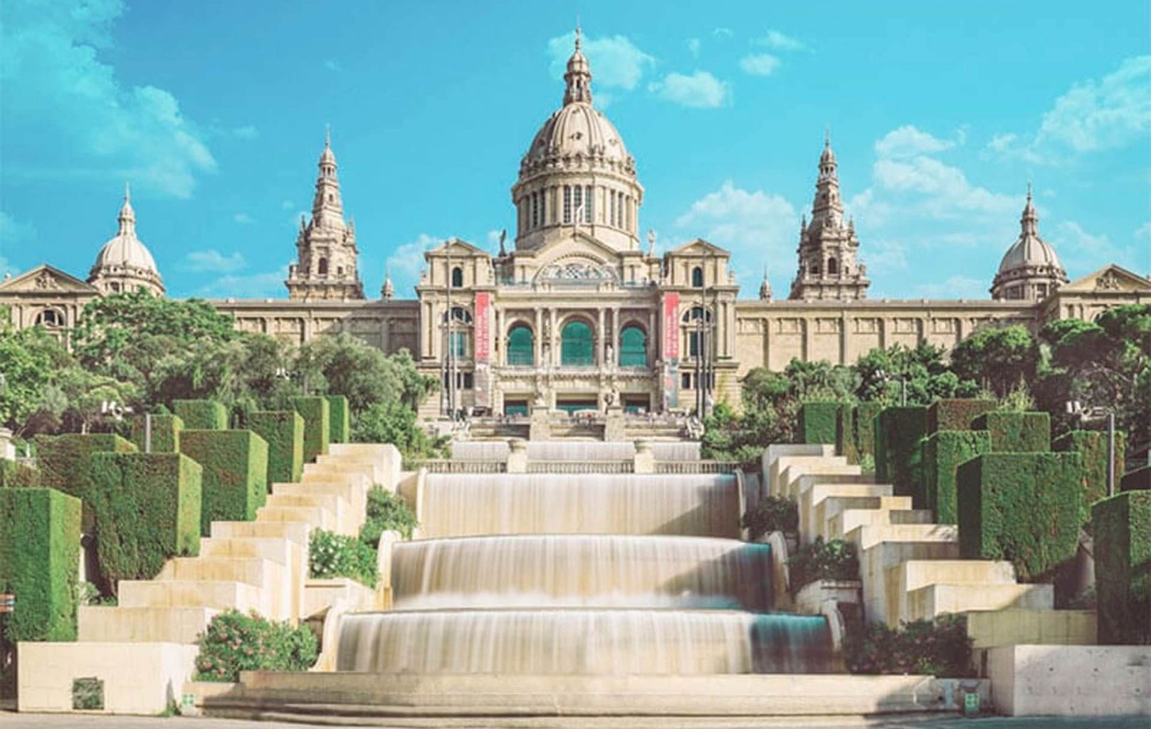 Un gran edifici històric amb una cúpula central i torres, el Museu Nacional d'Art de Catalunya (MNAC) presenta fonts en cascada i nivells i bardisses ben cuidades en primer pla sota un cel blau.