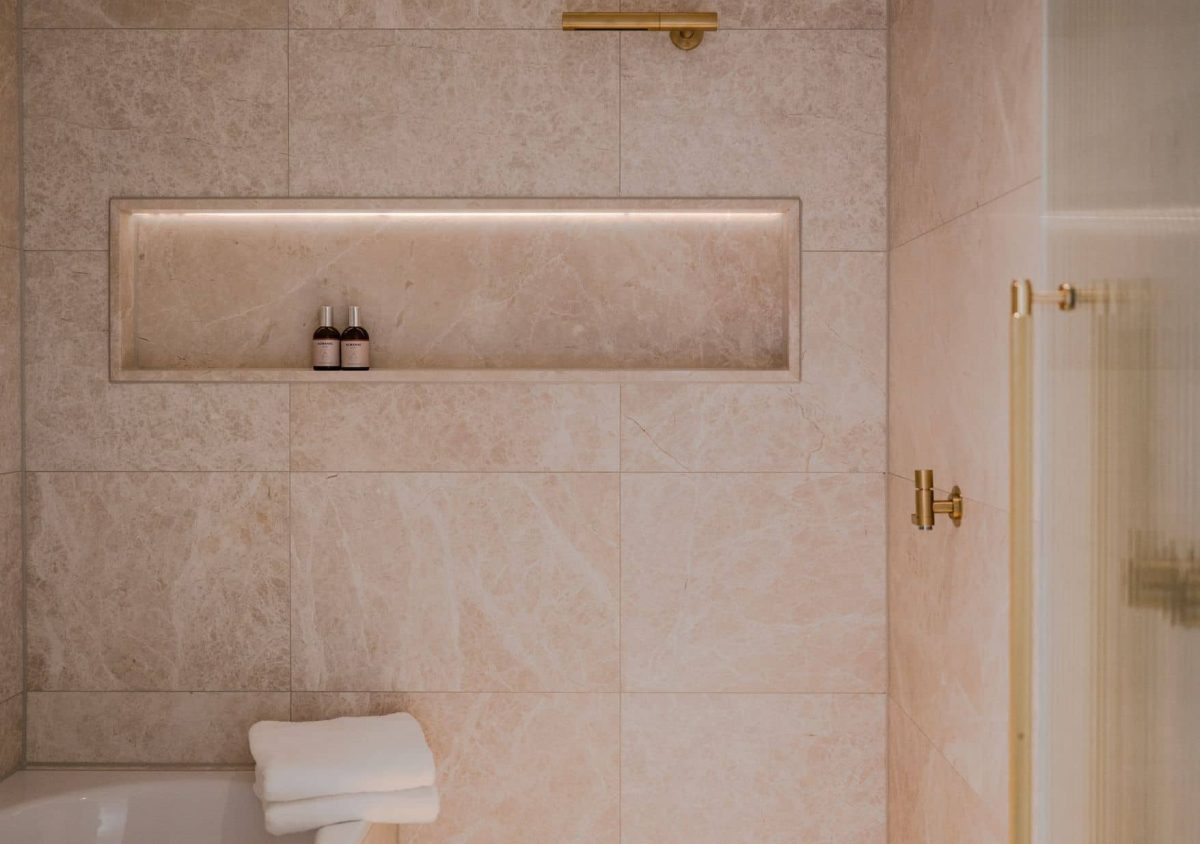 Ein modernes Zimmer-Badezimmer mit beigen Marmorfliesen, einem eingebauten Regal für zwei Flaschen, einem Handtuch auf dem Badewannenrand und schlichten goldenen Armaturen.