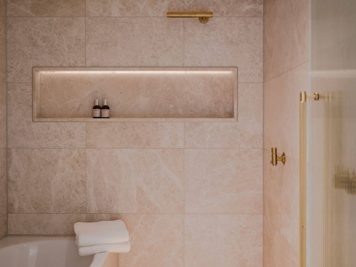 Ein modernes Zimmer-Badezimmer mit beigen Marmorfliesen, einem eingebauten Regal für zwei Flaschen, einem Handtuch auf dem Badewannenrand und schlichten goldenen Armaturen.