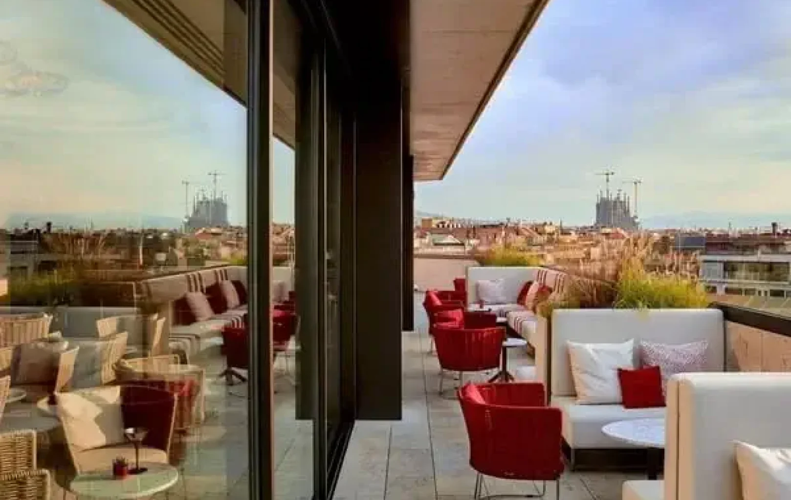 Terrassa exterior a la teulada amb seients encoixinats, cadires vermelles i taules petites —perfecta per a una Escapada de Tardor— amb vistes a un paisatge urbà amb grues i edificis distants sota un cel parcialment ennuvolat.