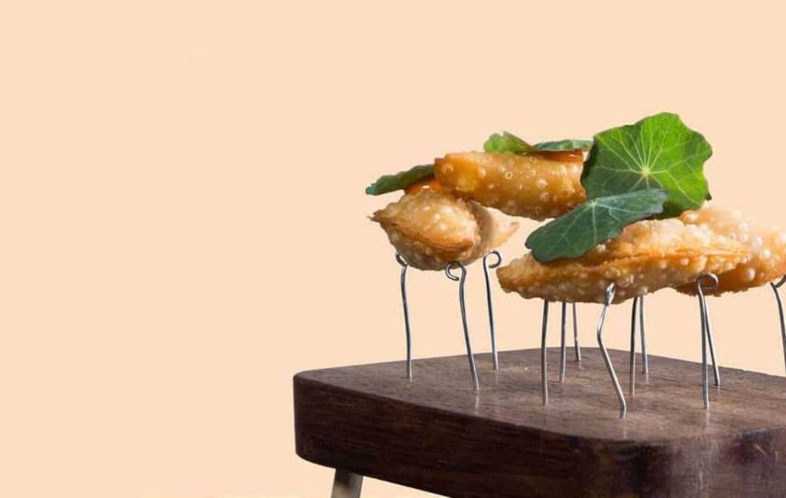 Tres albóndigas fritas se presentan en posición vertical sobre soportes de alambre en un bloque de madera, adornadas con hojas verdes, sobre un fondo beige liso: un bocado elegante inspirado en los elevados sabores del Azimuth Rooftop Bar.