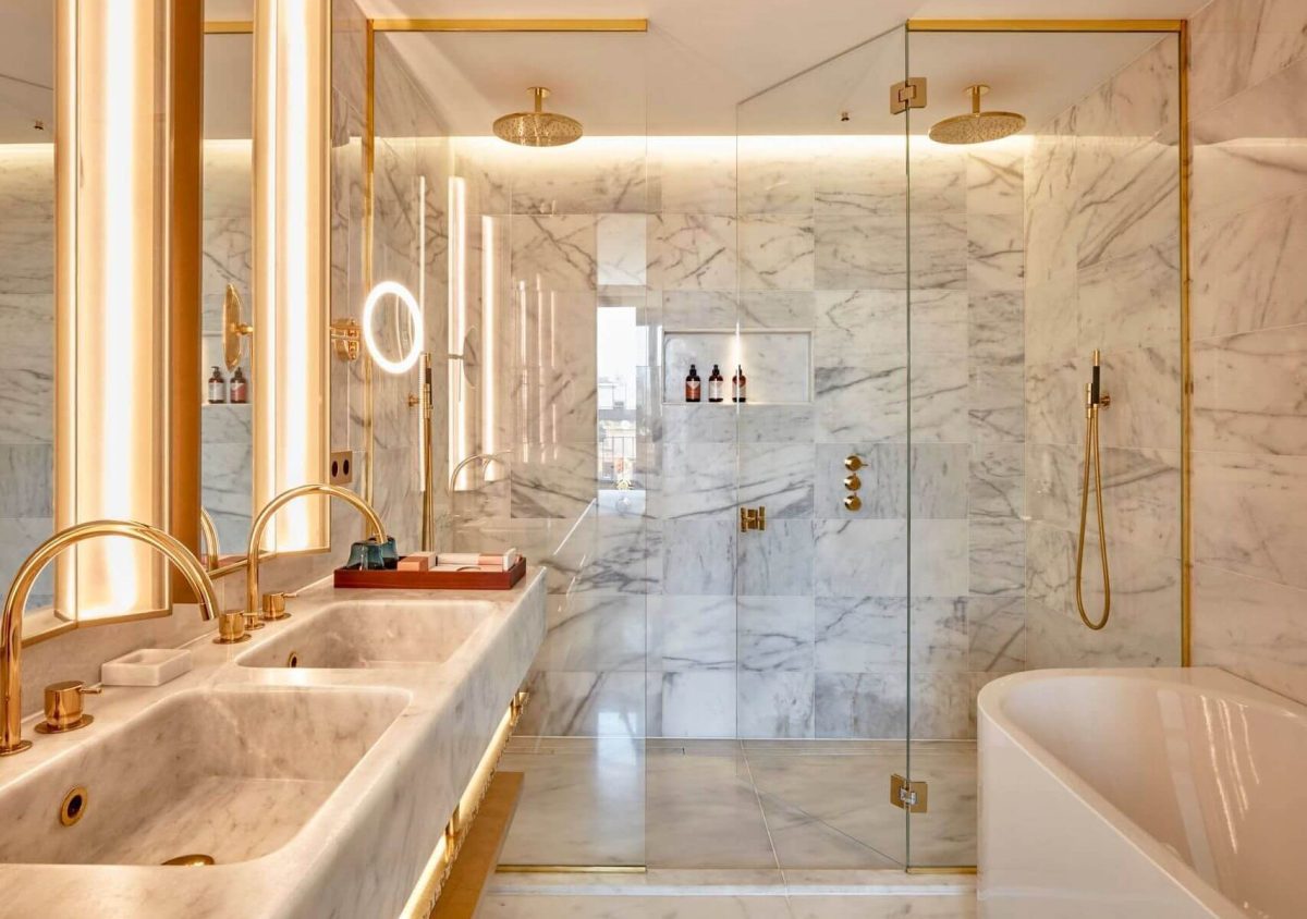Bany suite a l'Ć tic amb parets i terres de marbre, lavabo doble amb accessoris daurats, mampara de dutxa de vidre i banyera independent: un luxe perfecte per a dues habitacions.