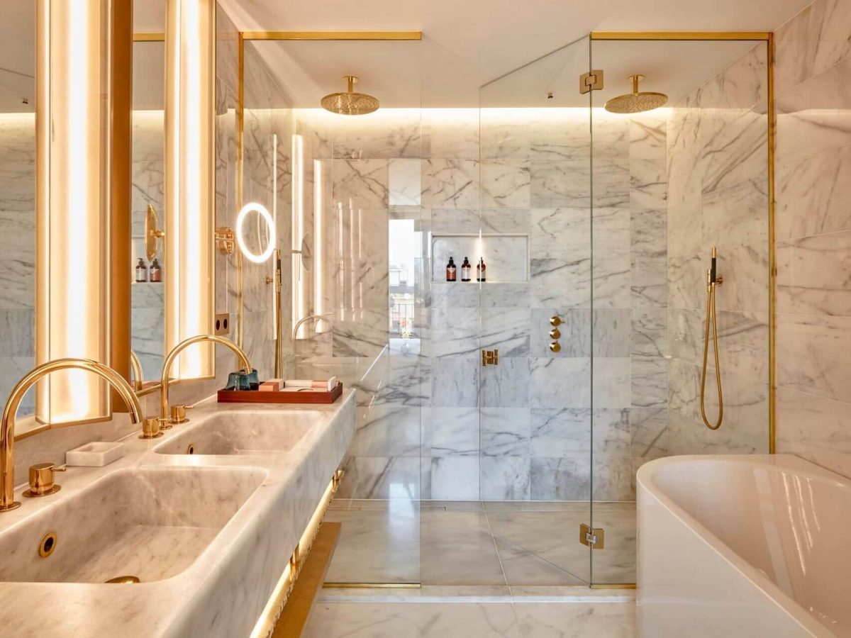 Luxuriöses Badezimmer in der Penthouse Suite mit Marmorwänden und -böden, Doppelwaschbecken, goldenen Armaturen, einer großen Glasdusche und einer freistehenden Badewanne - perfekt für einen Rückzugsort für zwei Schlafzimmer.