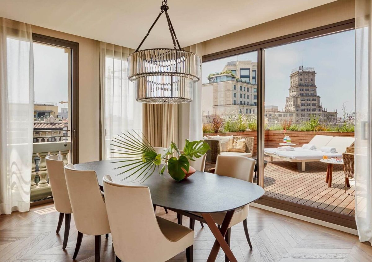 Una moderna zona de comedor con seis sillas se encuentra en la Suite Penthouse, junto a grandes ventanales que dan a una terraza con tumbonas y edificios de la ciudad visibles más allá de este elegante refugio de dos dormitorios.