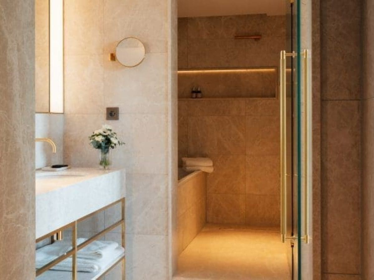 Modernes Badezimmer mit beigen Steinwänden, weißem Waschbecken, goldenen Armaturen, einem runden Spiegel und einer eingelassenen Badewanne mit gestapelten Handtüchern und Toilettenartikeln - perfekt, um sich in der Eleganz der Klimt Suite zu entspannen.