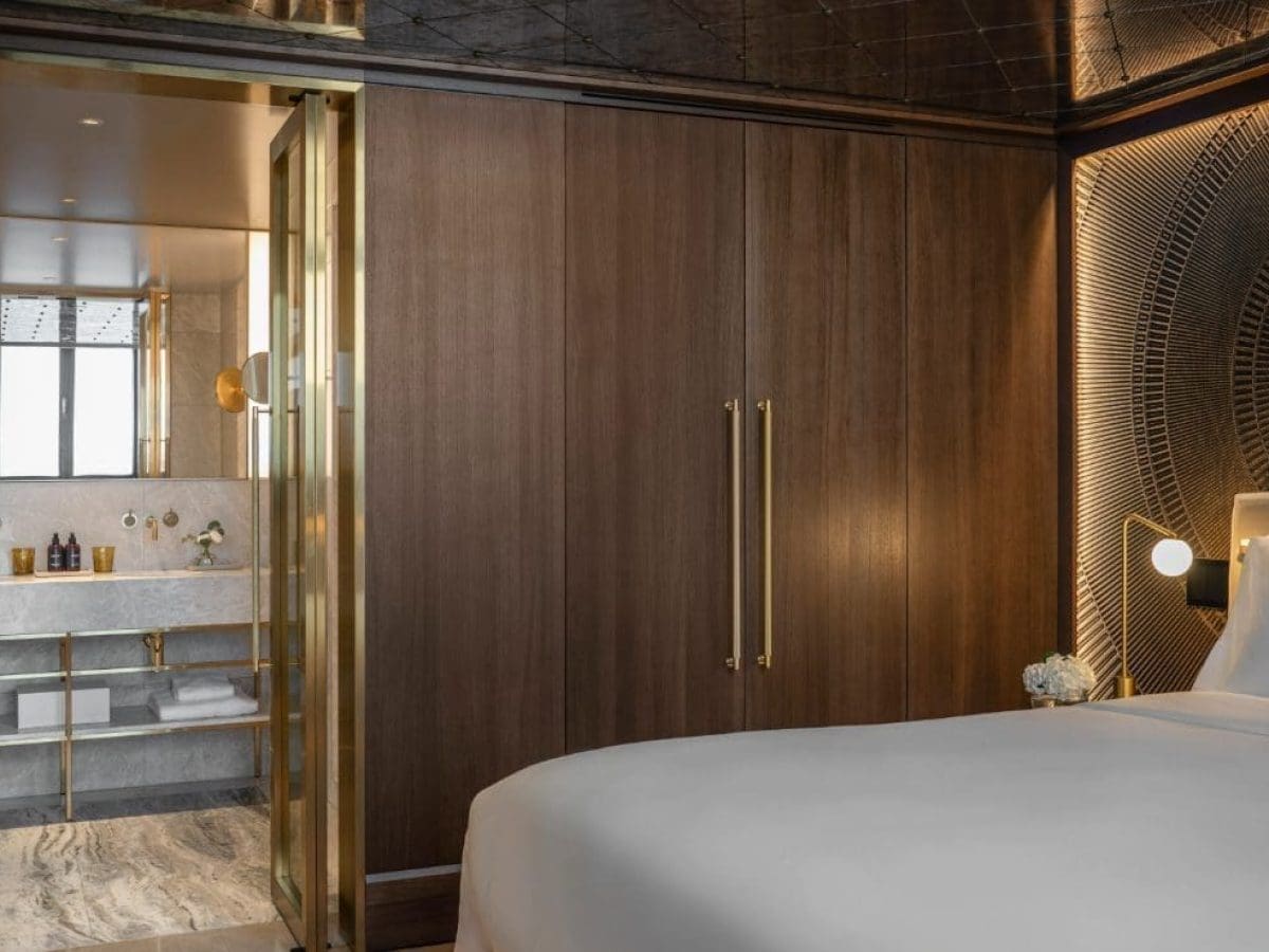 Moderne Klimt-Suite mit großem Bett, holzgetäfelten Wänden, goldenen Akzenten und einem offenen Blick in ein Marmorbad mit Doppelwaschbecken.