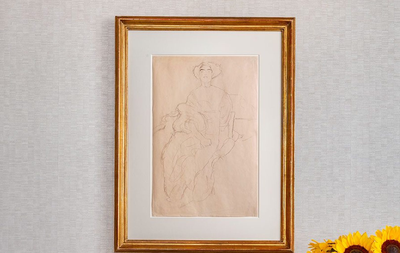 A gold-framed sketch of a seated person is hung on a light textured wall, with sunflowers partially visible in the lower right corner.