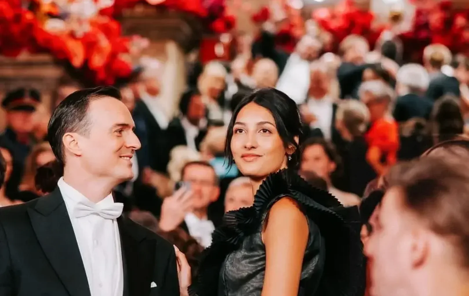 A woman in a black dress and a man in a tuxedo stand at a formal event, capturing the elegance of a Night At The Ball in the midst of a crowded, sophisticated ballroom.