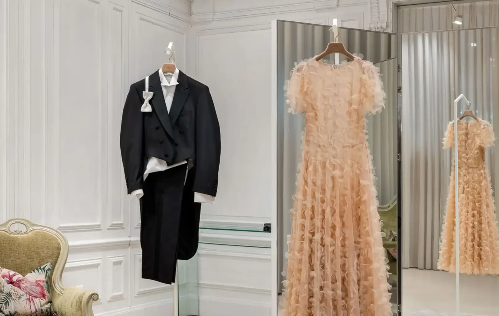A black tuxedo and a light peach ruffled gown, perfect for A Night At The Ball, are displayed on hangers in an elegant, white-paneled fitting room with a mirror and plush chair.