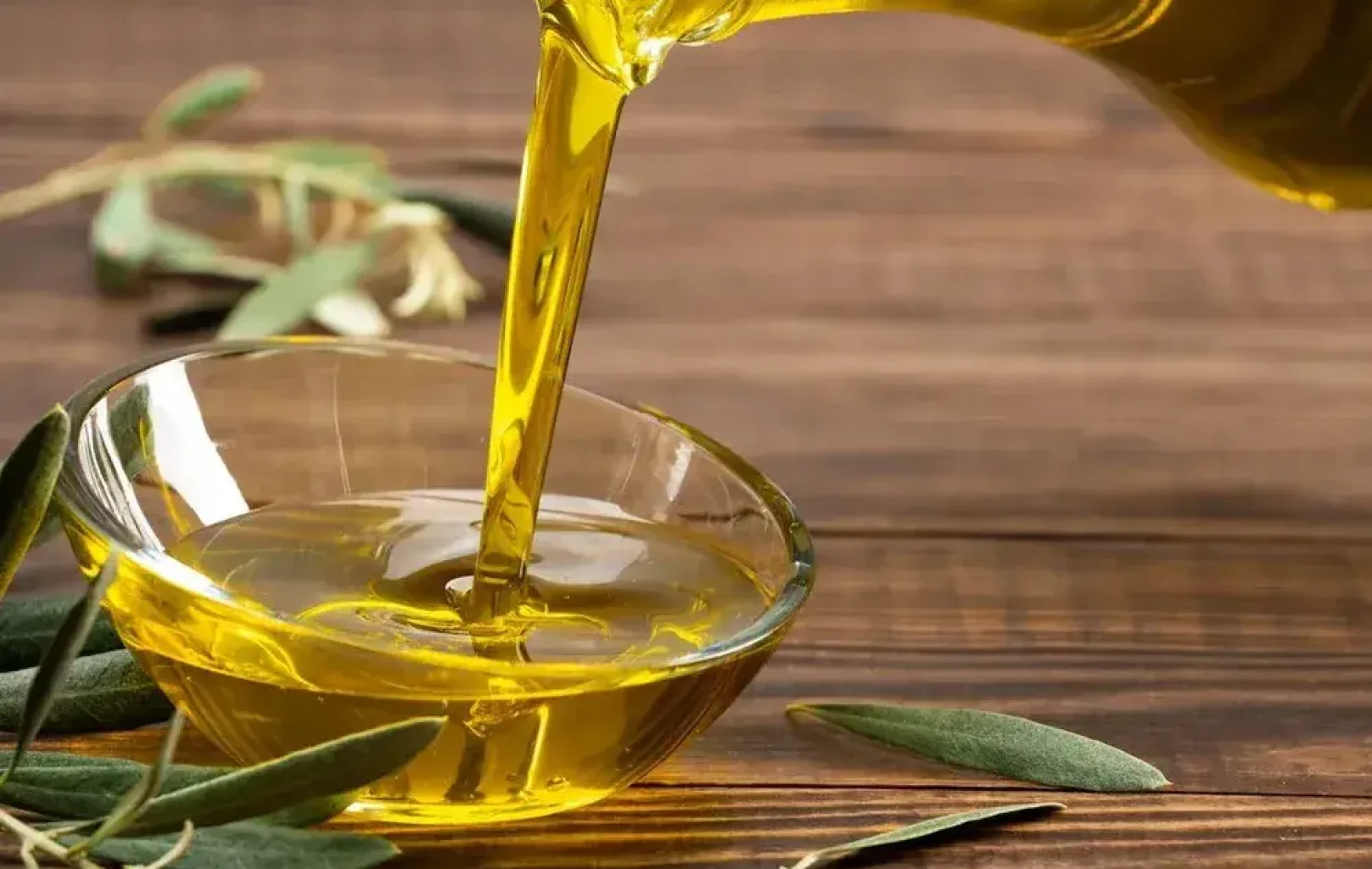 Olive oil being poured from a glass bottle into a small glass bowl, with olive branches on a wooden surface—an inviting scene that highlights how to make olive oil from scratch.